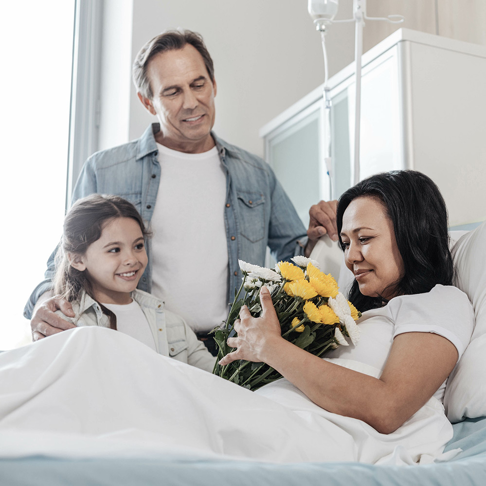 Mother holding flowers being visited by husband and daughter in hospital